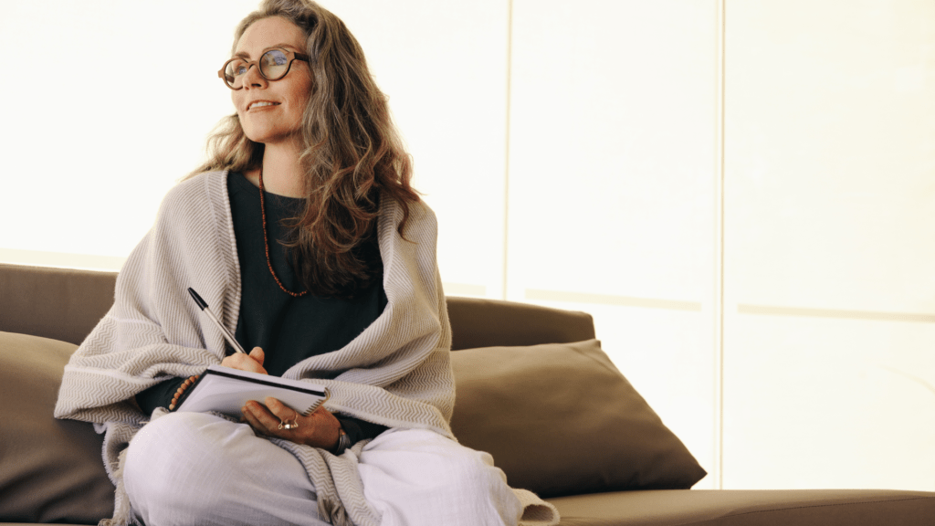 A woman enhancing emotional awareness through reflective journaling, sitting by a window with a serene expression, promoting mindfulness and self-discovery.